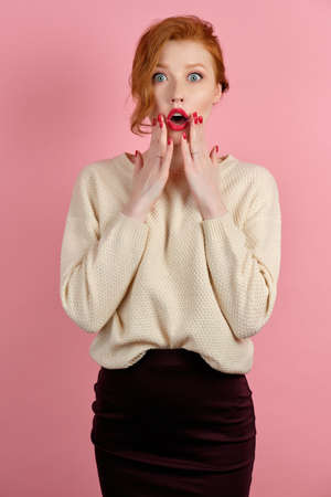 Redhead girl with red lipstick and red manicure looks shocked at the camera, raising her hands to her faceの写真素材