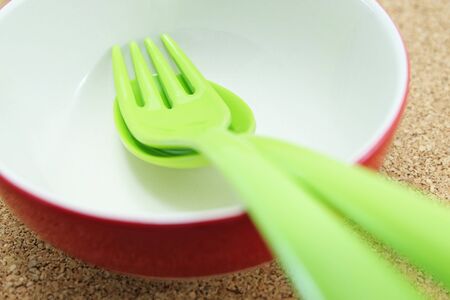green plastic spoon and fork are placed in a red bowl on cork plywood boardの写真素材