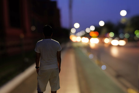 Backside of Man who stands on street corridor and looks forward to the road and some vehicles at night in blur modeの写真素材
