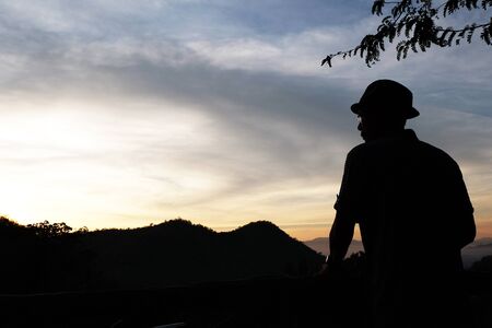 a man who wears a hat,an eye glasses Selected focus with some mountains,tree and gradient light orange-blue sky while before sunrise silhouette modeの写真素材