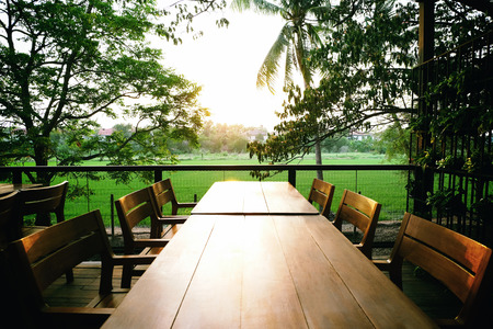 Sun shining from the center of frame to long wooden table
 with six wooden chairs ,metal fence
and Rice fields backgroundの写真素材