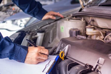 Hands of young professional mechanic in uniform writing on clipboard against car in open hood at the repair garage. Maintenance se. car service and maintenanceの写真素材