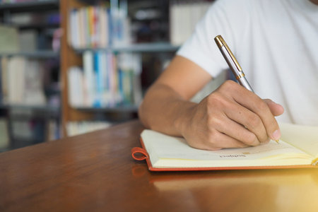 Students sit in the library. Holding a pen is writing on a notebook.