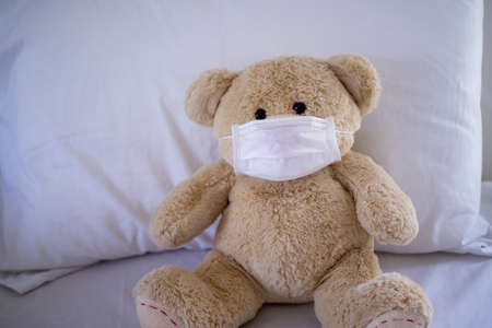 The teddy bear is sitting on a bed wearing a mask to protect against germs and viruses. Hygiene for children conceptの写真素材
