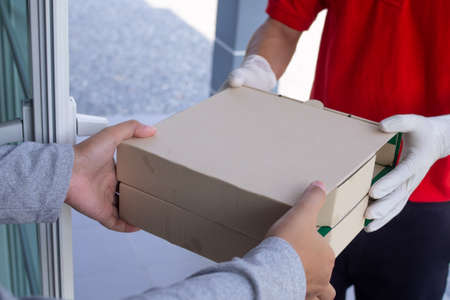 A food deliveryman wearing a red uniform and sanitary gloves, delivering a food box to a customer is calls online to eat at home. online delivery concepts.の写真素材