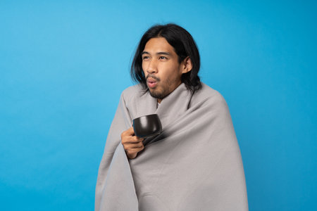 The young man was sick and coughed with a warm cup of water and wrapped in a cloth against a blue background. The patient is sick with runny nose and chills. Influenza patient conceptの写真素材