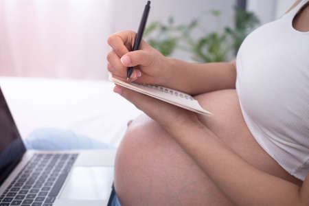 A pregnant woman works from home during her birth. A woman holding a notebook and a pen to take information from the computerの写真素材
