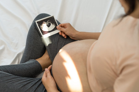 A pregnant woman is looking at an ultrasound photo of fetus. Mother gently touches the baby on...