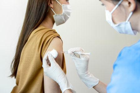 Asian woman wearing a medical mask receives coronavirus vaccine from a doctor. People are vaccinated against virus to prevent infection with the virus and stop its spread.の写真素材