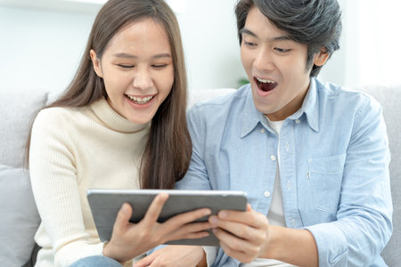 Asian couple smiling and happily use smartphone to online shopping. Husband ready to pay birthday present for beautiful wife. Find accommodation online for honeymoon, App online, book flight tickets.の写真素材