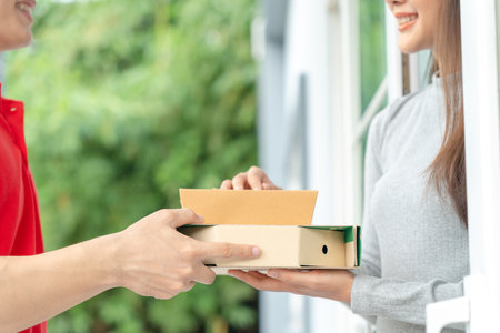 Happy smiling Asian woman receives paper box parcel of food from courier front house. Delivery man send deliver express. online shopping, paper container, takeaway, postman, delivery service, packagesの写真素材