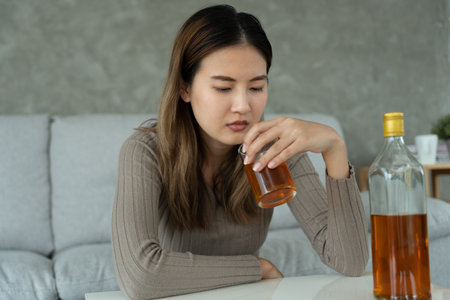 Depressed young Asian woman addicted feeling bad drinking whiskey alone at home, stressed frustrated lonely drinking alcohol suffers from problematic liquor, alcoholism, life and family problemsの写真素材
