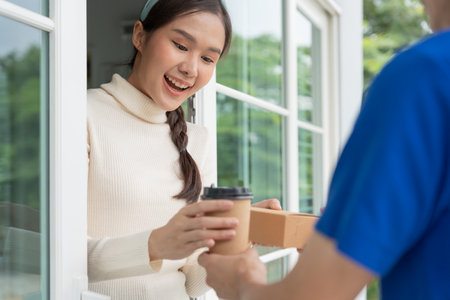 Happy smiling Asian woman receives paper box parcel of food from courier front house. Delivery man send deliver express. online shopping, paper container, takeaway, postman, delivery service, packagesの写真素材
