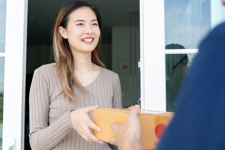 Happy smiling woman receives boxes parcel from courier in front house. Delivery man send deliver express. online shopping, paper containers, takeaway, postman, delivery service, packagesの写真素材