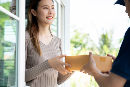 Happy smiling woman receives boxes parcel from courier in front house. Delivery man send deliver express. online shopping, paper containers, takeaway, postman, delivery service, packagesの写真素材