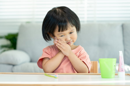 little asian girl presses hand to cheek, suffers from pain in tooth. Teeth decay, dental problems, child emotions and facial expression, oral health care, reducing sweets, fluorine coatingの写真素材