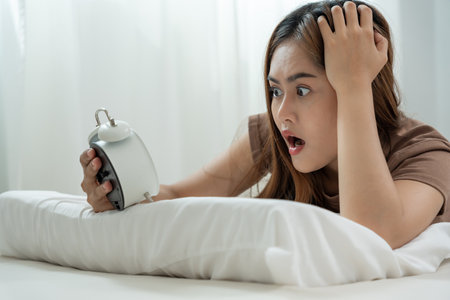 Shocked Asia young woman looking at clock alarm, lady is late appointment or work or class study, overslept, difficulty get up in mornings, time worried, not on time, sleepy, over due, Hurry up timeの写真素材