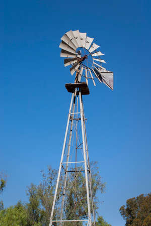 ranch aluminum vintage windmill with blue sky in the background and trees on the bottom. Room for textの写真素材