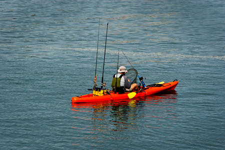 orange kayak, yellow paddles, with fishing poles in the back. Man with his back to the camera holding a net getting ready to pull up a fish.の写真素材