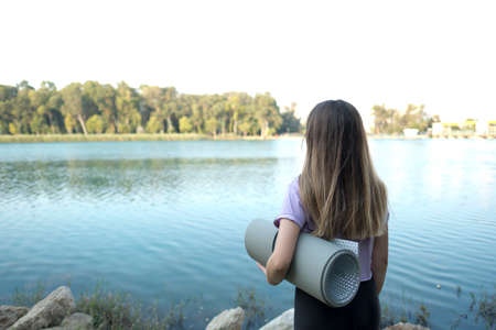 Girl in sport clothes holding a yoga mat, looking at river.