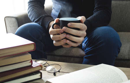 Young man is sitting on a sofa and reading a book while holding a cup of coffee.