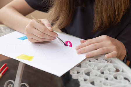 Teenager's hands painting with fuchsia watercolor and brush on a color wheel diagram on white paper. Close-up view on a glass table outdoors. Copy space. Concept of artistic learning and creative expressionの写真素材
