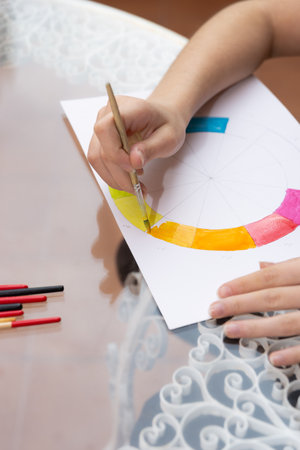 Teen hand painting a color wheel with yellow watercolor on a white sheet. Top-down view. School supplies on a decorative white metal table. Copy space. Concept of educational practice and artistic developmentの写真素材