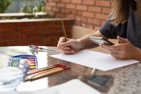 Student girl using smartphone while drawing on white paper, school tools on outdoor table. Copy...