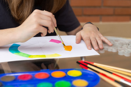 Young hands apply orange watercolor on a partially painted color wheel. School supplies and watercolor set on a glass table outdoors. Close-up view. Concept of artistic education and creative developmentの写真素材