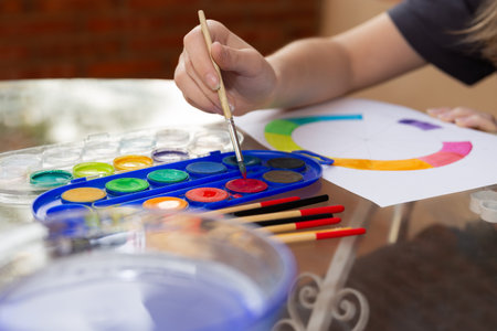 Close-up of child's hand picking up red watercolor with a paintbrush from a palette. Blurred background with half-painted color wheel. Outdoor scene. Copy space.の写真素材