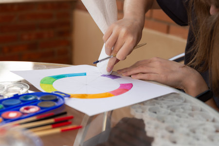 Teenage girl's hands wiping excess watercolor from a painted color wheel using white paper towel. Outdoor glass table with school supplies. Copy space.の写真素材