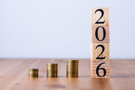 Vertical tower of wooden blocks with number 2026 next to golden coin piles on wooden table and gray...