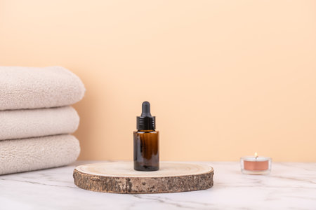 Amber glass dropper bottle on rustic wooden base with beige towels and candle on marble table against peach background. Copy space. Concept of self care and natural beautyの写真素材