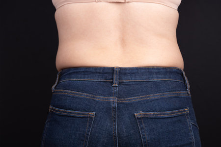Rear view of a woman's torso wearing tight jeans and a bra, black background. Copy space. Concept of real curves, self-esteem, and body diversityの写真素材