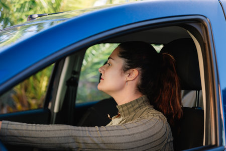 Young woman driving a blue car with a focused expression and upright posture, side portrait in an...