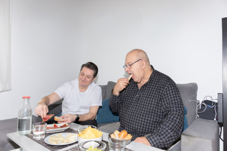 Mature male couple sharing snacks and conversation in the living room, natural and spontaneous...