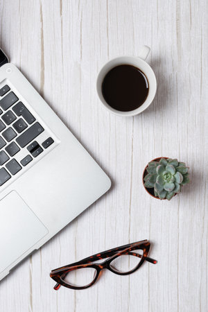 Top vertical view of laptop, coffee cup, glasses and succulent on light wood background, natural...