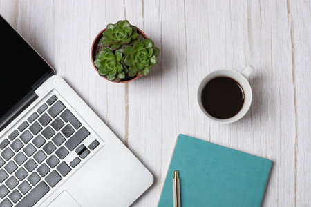 Top horizontal view of laptop, coffee cup, succulents and turquoise notebook on light wooden...