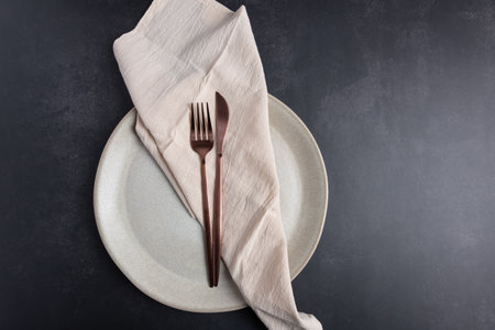 Horizontal flat lay of light ceramic plate with copper cutlery and beige linen napkin on dark gray...