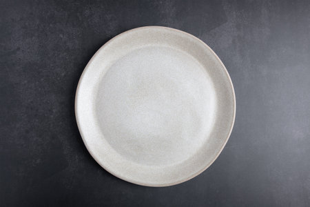 Empty beige ceramic plate on dark background with copy space, top view. Concept of minimalist...