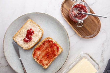 Top view of toast with butter and strawberry jam on a gray plate, with knife, butter dish, and jam jar with spoon on white marble. Concept of homemade breakfast and bright kitchenの写真素材