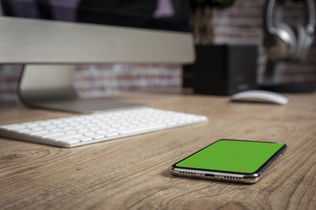 full screen Smartphone on the top of a wooden table in front of a office. Green screen for video, copy space.の写真素材
