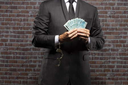 Brazilian man holding bills of money with a handcuff in a jail. concept of corruption, corrupt politicians, illegal businesses. brick background.の写真素材