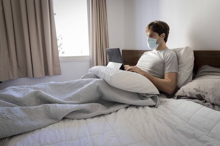 Man working in bed, doing home office because he is sick. using a mask over the mouth and nose.の写真素材