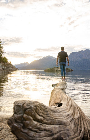 Man standing on a tree trunk and looking at a lake at sunset.の写真素材