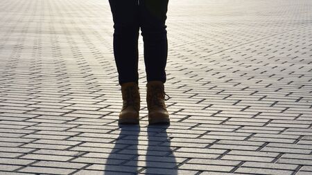 A girl in boots is standing on the pavement in the evening light. A woman stands alone in the middle...