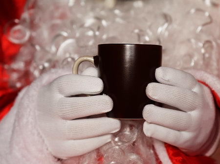 Santa Claus holding hands in a dark brown mug of tea in front of a white beardの写真素材
