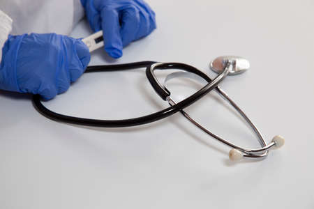There is a stethoscope on a white table. On the white surface is a stethoscope and the hands of a physician in gloves with a thermometer.の写真素材