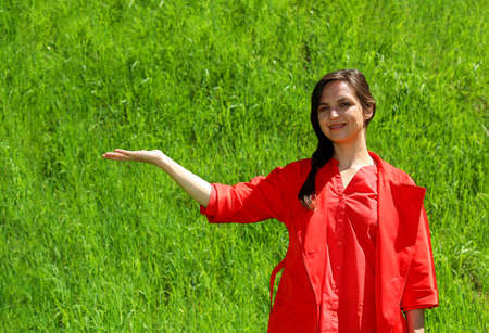 A smiling girl in red clothes on a background of green grass, a hand gesture in the side. A young, confident woman in the background of the green lawn raised her hand to the side. Space for advertisinの写真素材