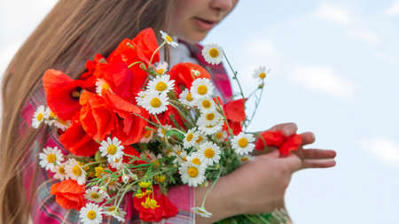 The girl stands against the background of wild flowers and holds a bouquet of poppies and daisies in her hands. A girl in the field was collecting a bouquet of wild flowers.の写真素材